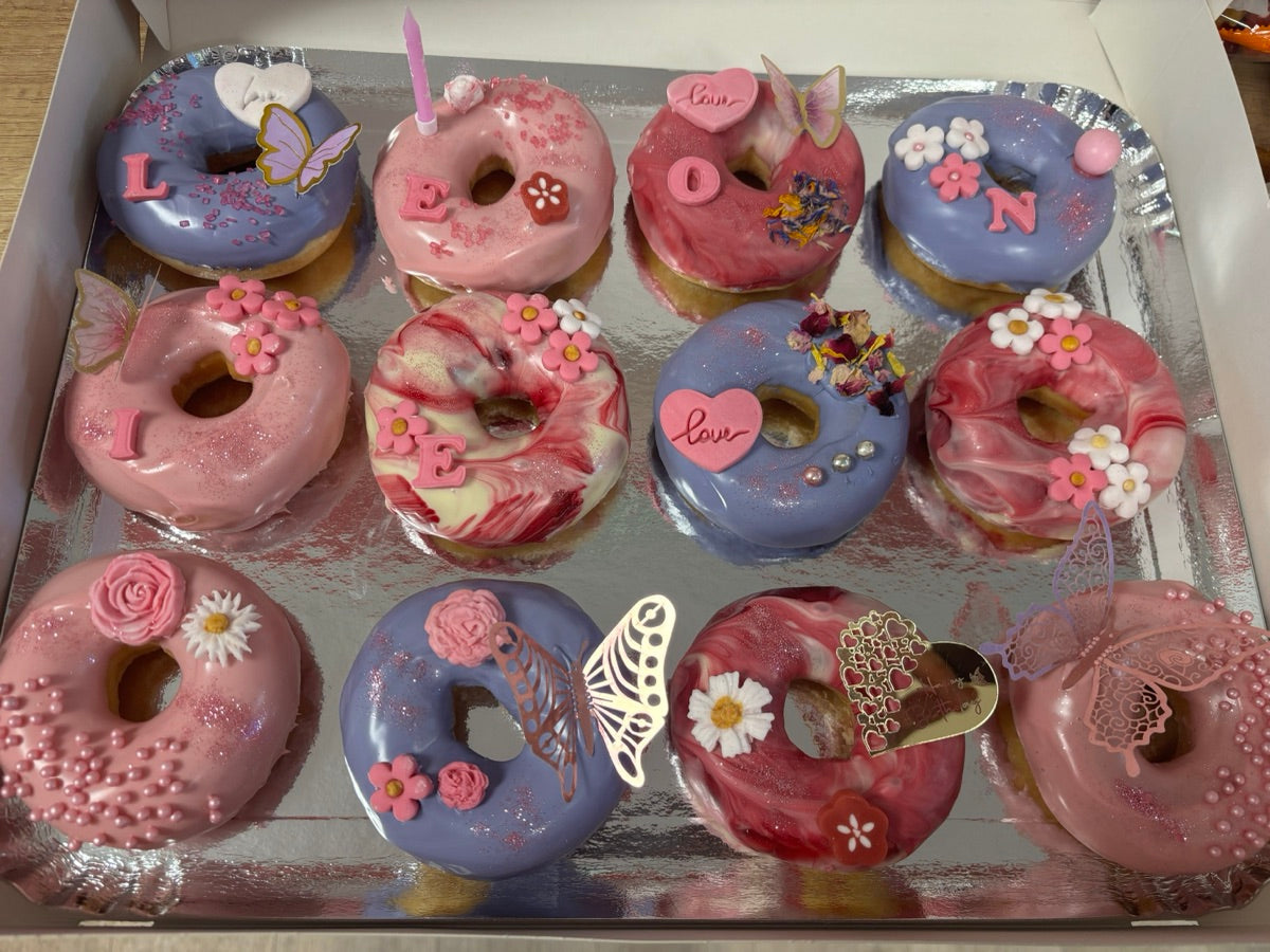 Des glaçages roses et lilas, des petites fleurs, des papillons et des lettres : un plateau de donuts décorés pour faire la fête.