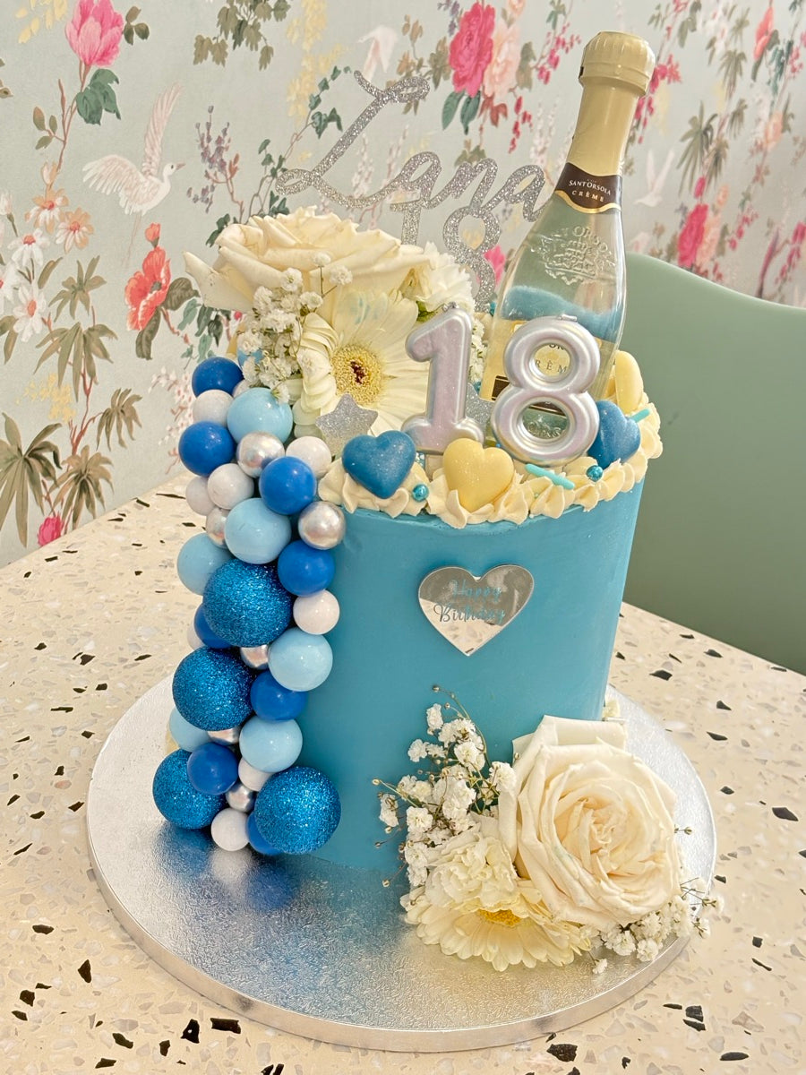 Un layer cake bleu décoré de roses blanches, d’une cascade de sphères et de décors bouteille et chiffres argentés.
