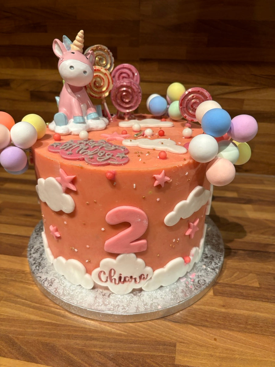 Un gâteau rose avec une licorne, des nuages blancs, des étoiles et des boules multicolores pour un anniversaire plein de fantaisie.